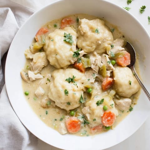 A close-up of rustic Chicken and Dumplings in a white bowl, garnished with fresh parsley and served alongside a spoon for a comforting meal.
