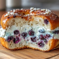A golden-brown Blueberry Burst Protein Bagel topped with creamy Greek yogurt frosting, dotted with juicy blueberries and ready for a healthy breakfast.  